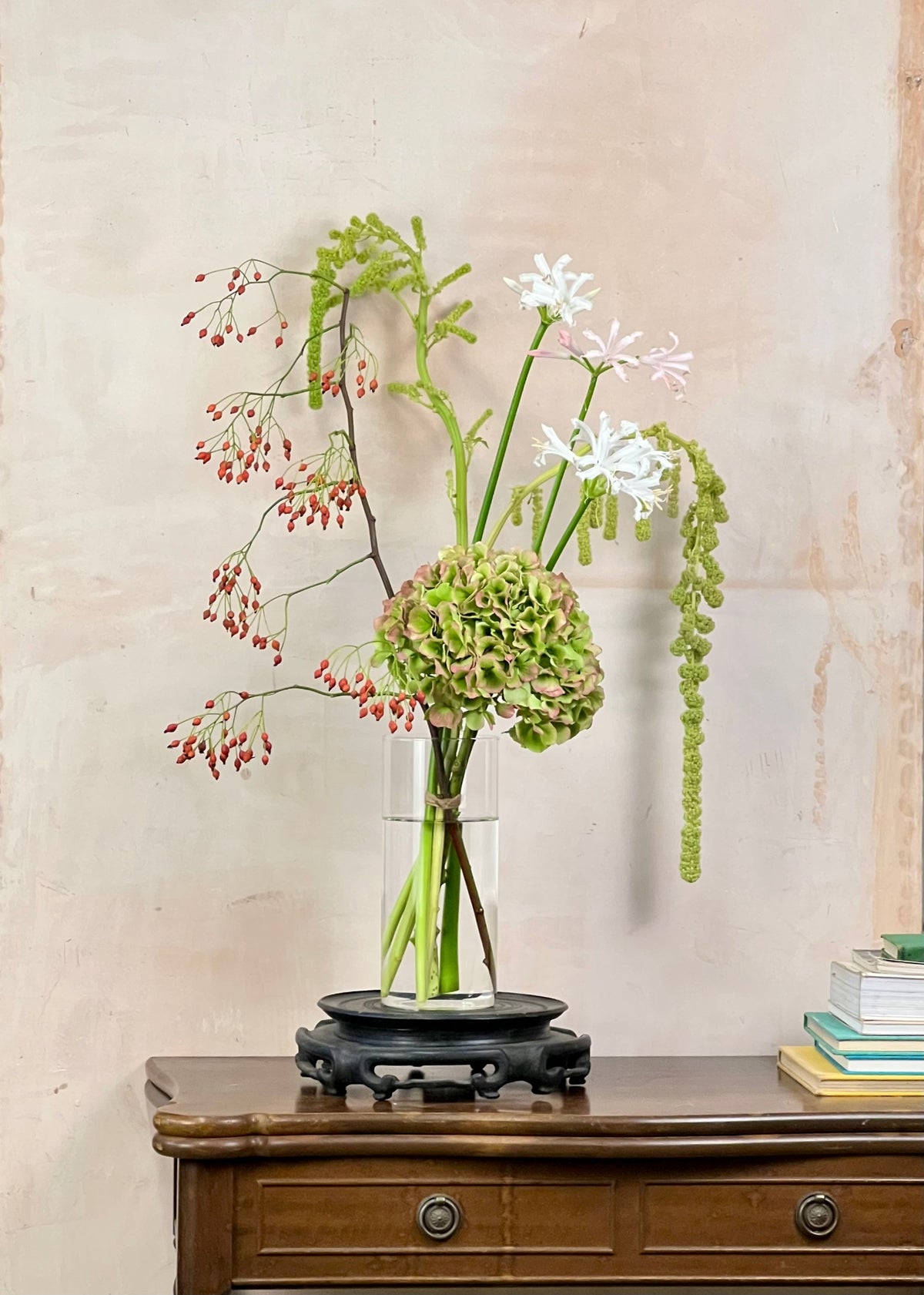 A floral arrangement in a clear glass vase with a black saucer, displayed on a wooden console table against a textured beige wall.