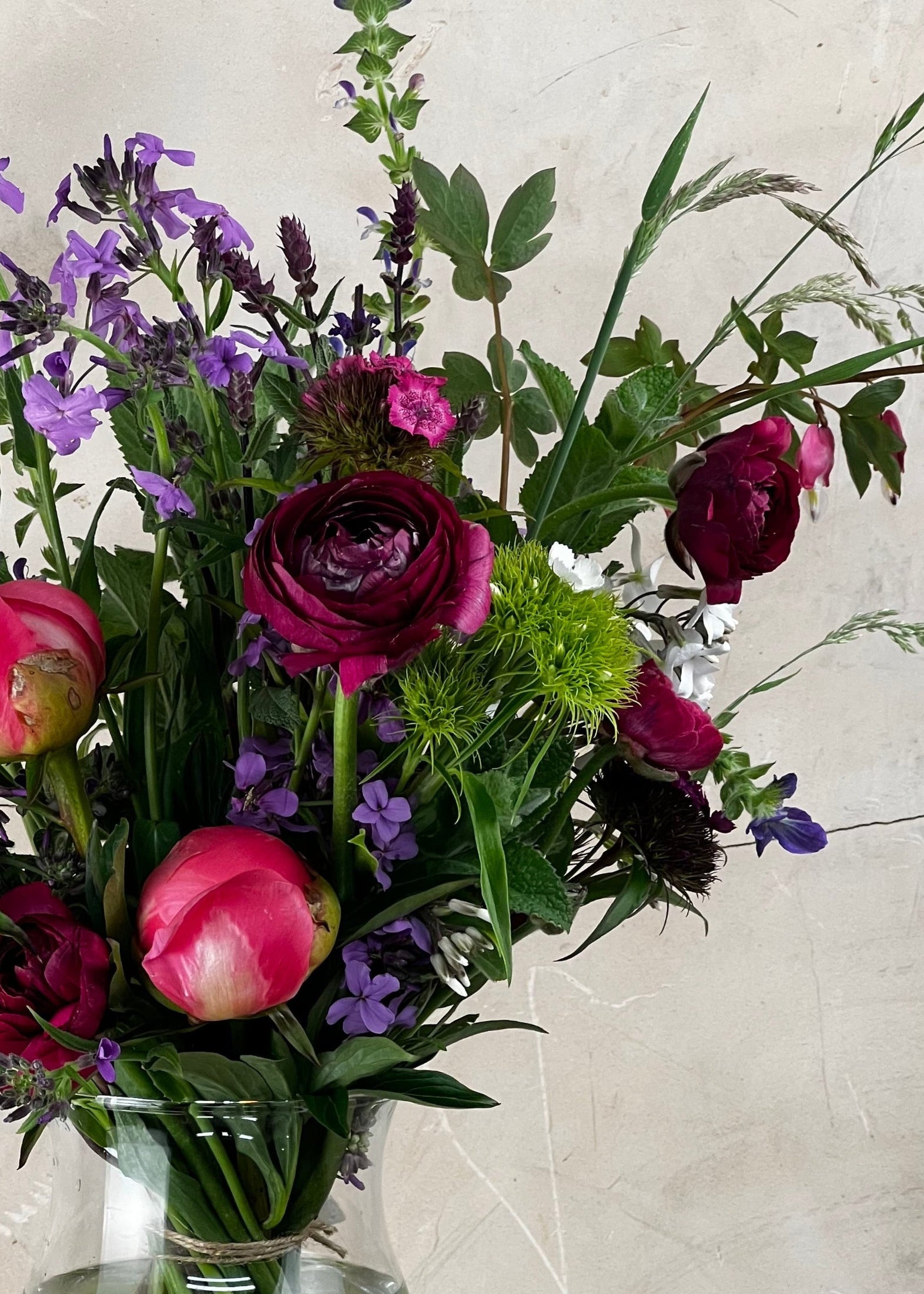 Bouquet of flowers with pink tulips, purple and red ranunculus, and greenery in a clear vase against a light background.