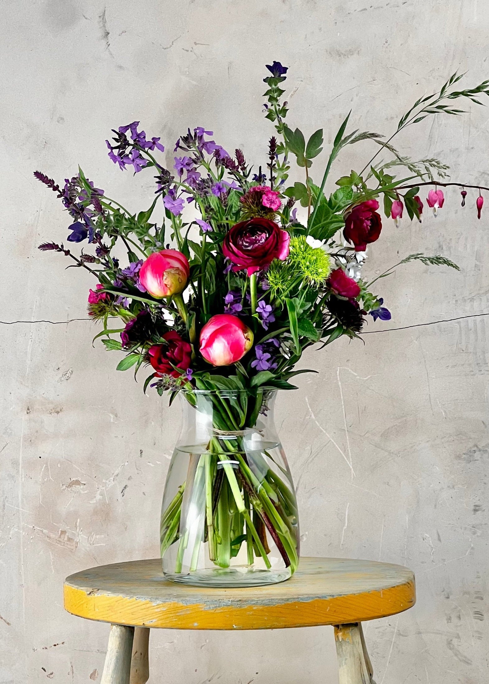 Bouquet of flowers in a glass vase on a wooden stool against a concrete wall.