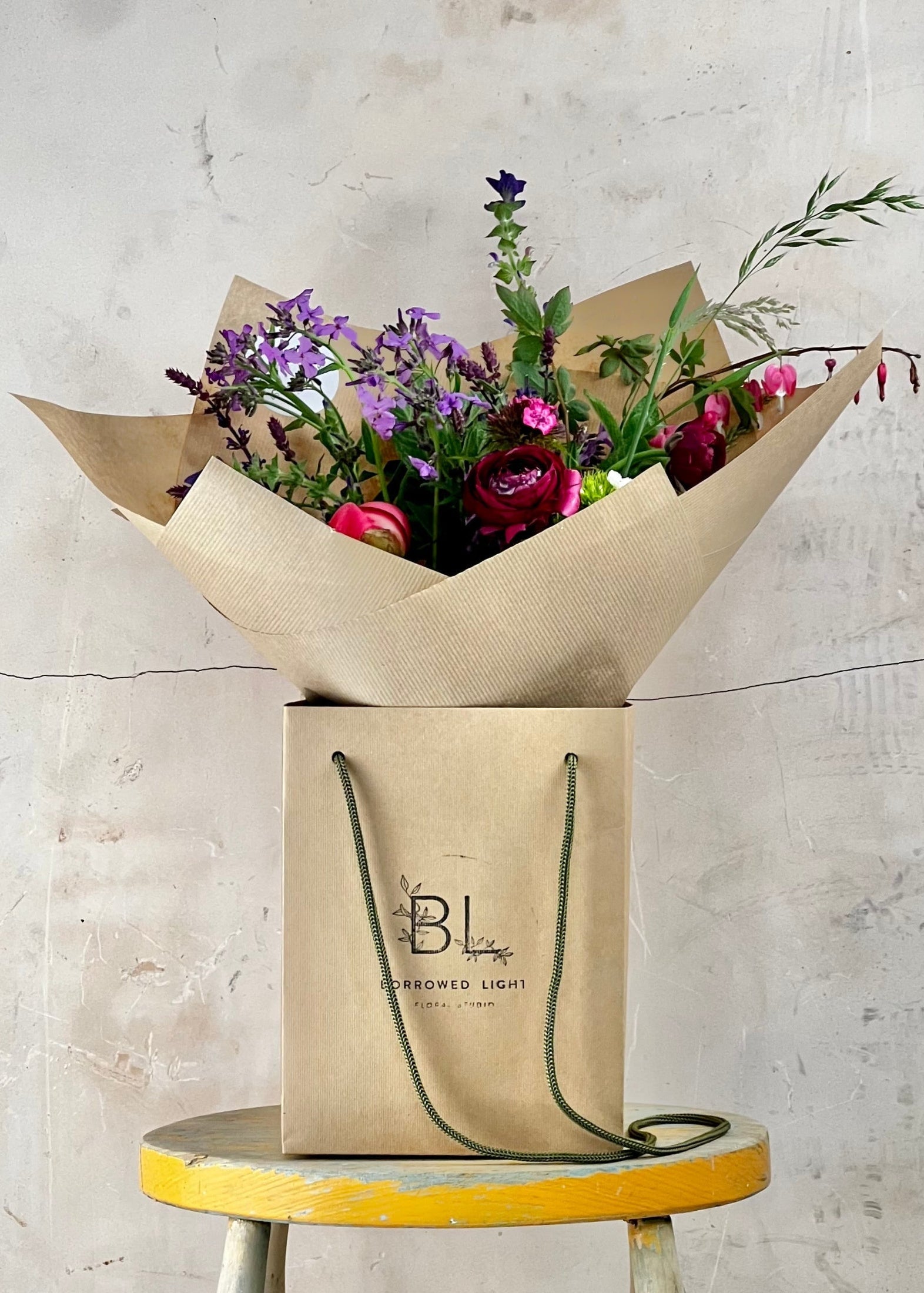Floral arrangement in a brown paper bag on a small stool against a textured wall.