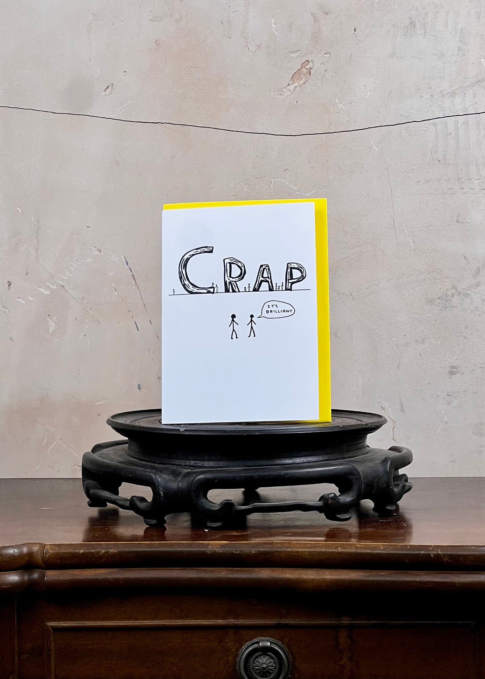 Card with 'Crap' text and illustration on a stand against a textured wall.