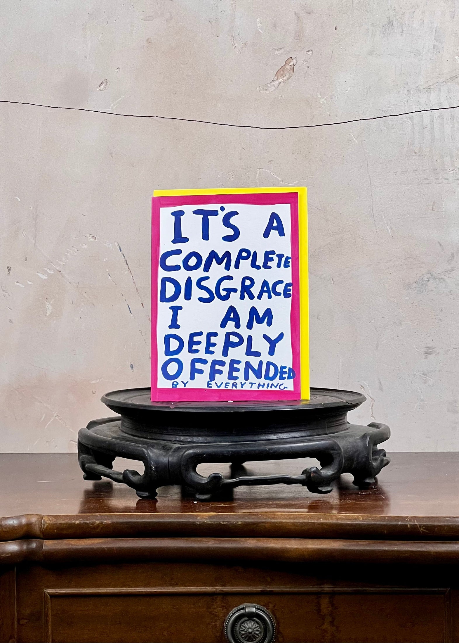 Card with text 'It's a complete disgrace I am deeply offended' on a stand against a textured wall.