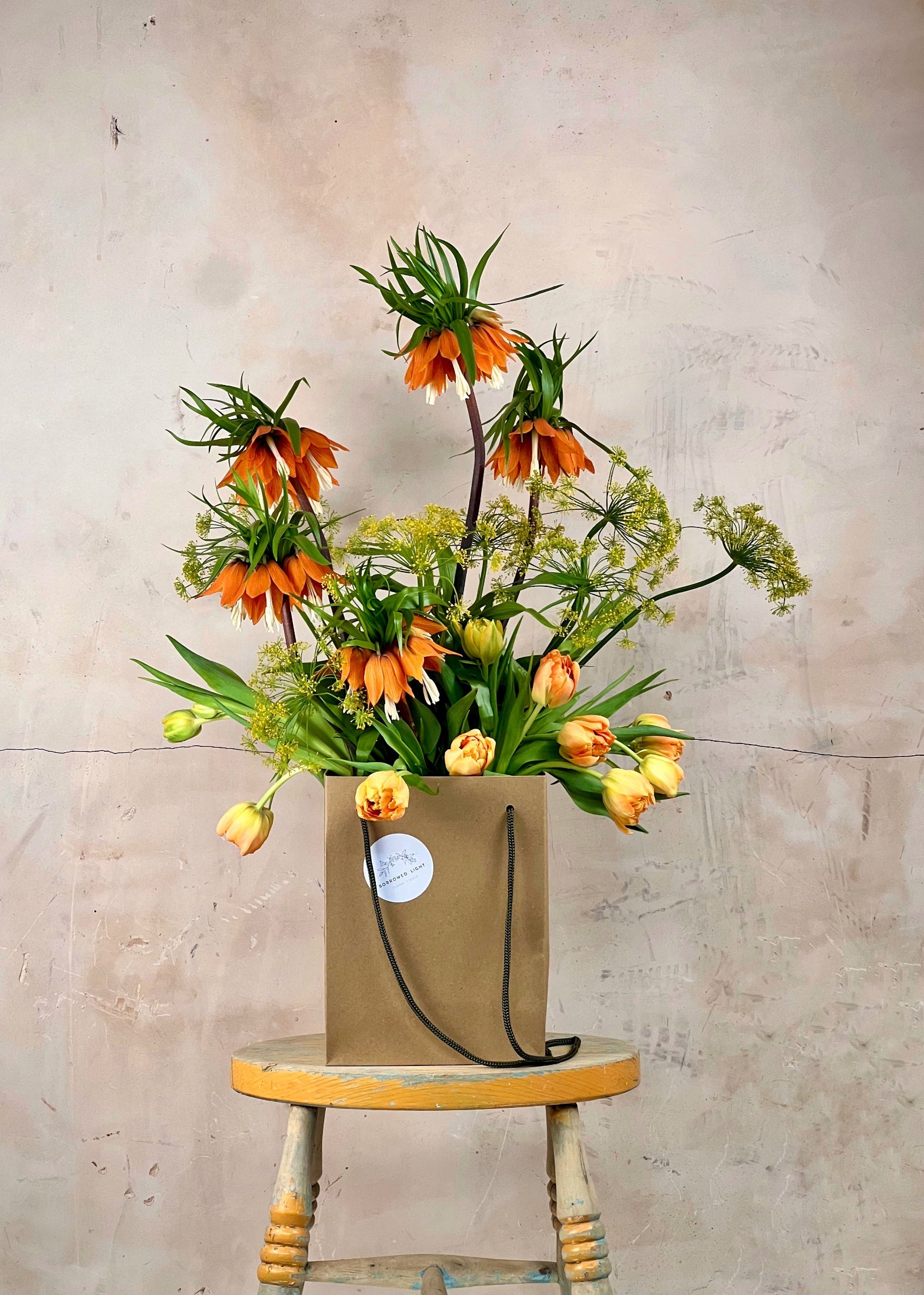 Floral arrangement in a brown paper bag on a wooden stool against a beige wall.
