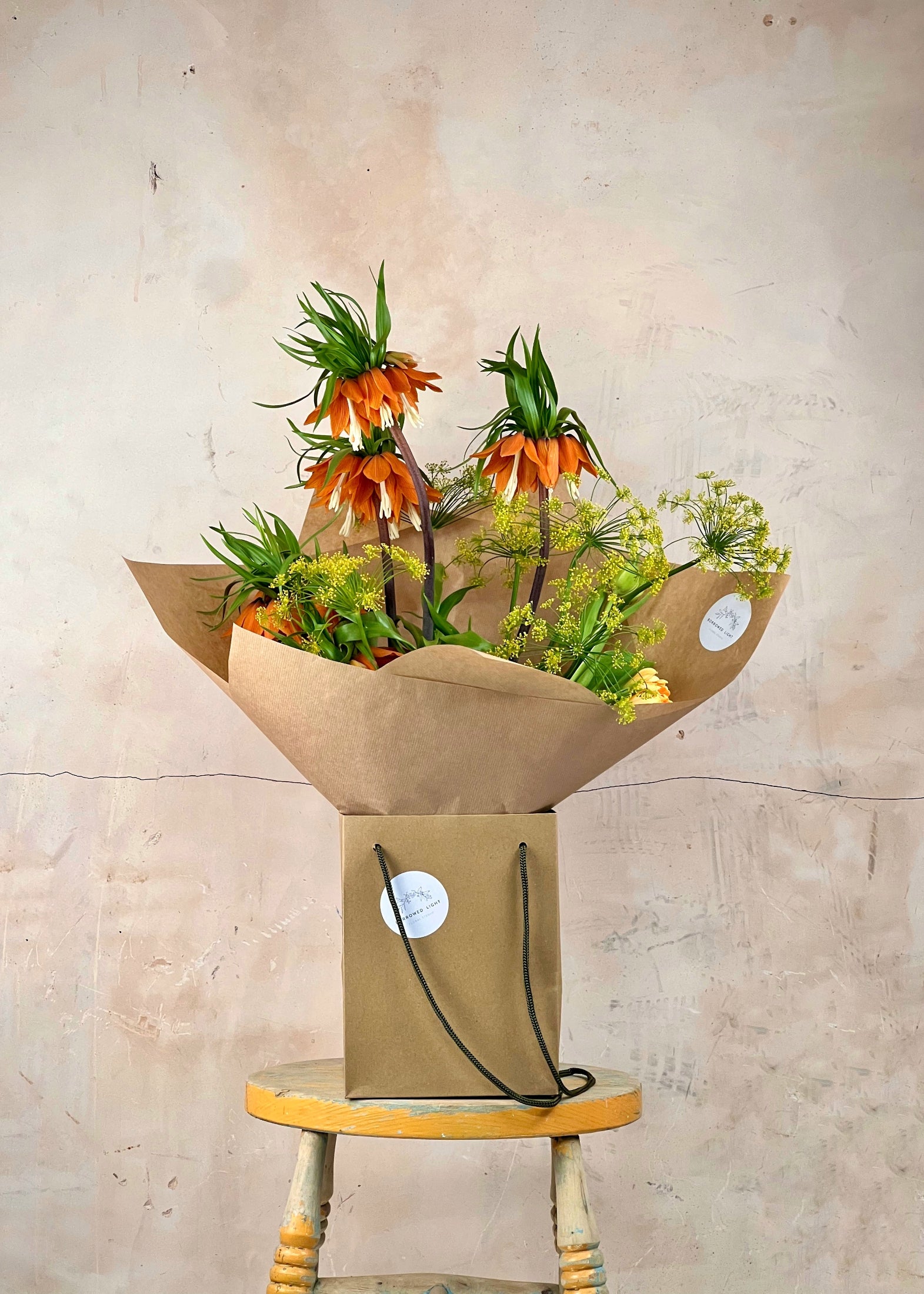 Bouquet of flowers in brown paper packaging on a wooden stool against a beige wall.