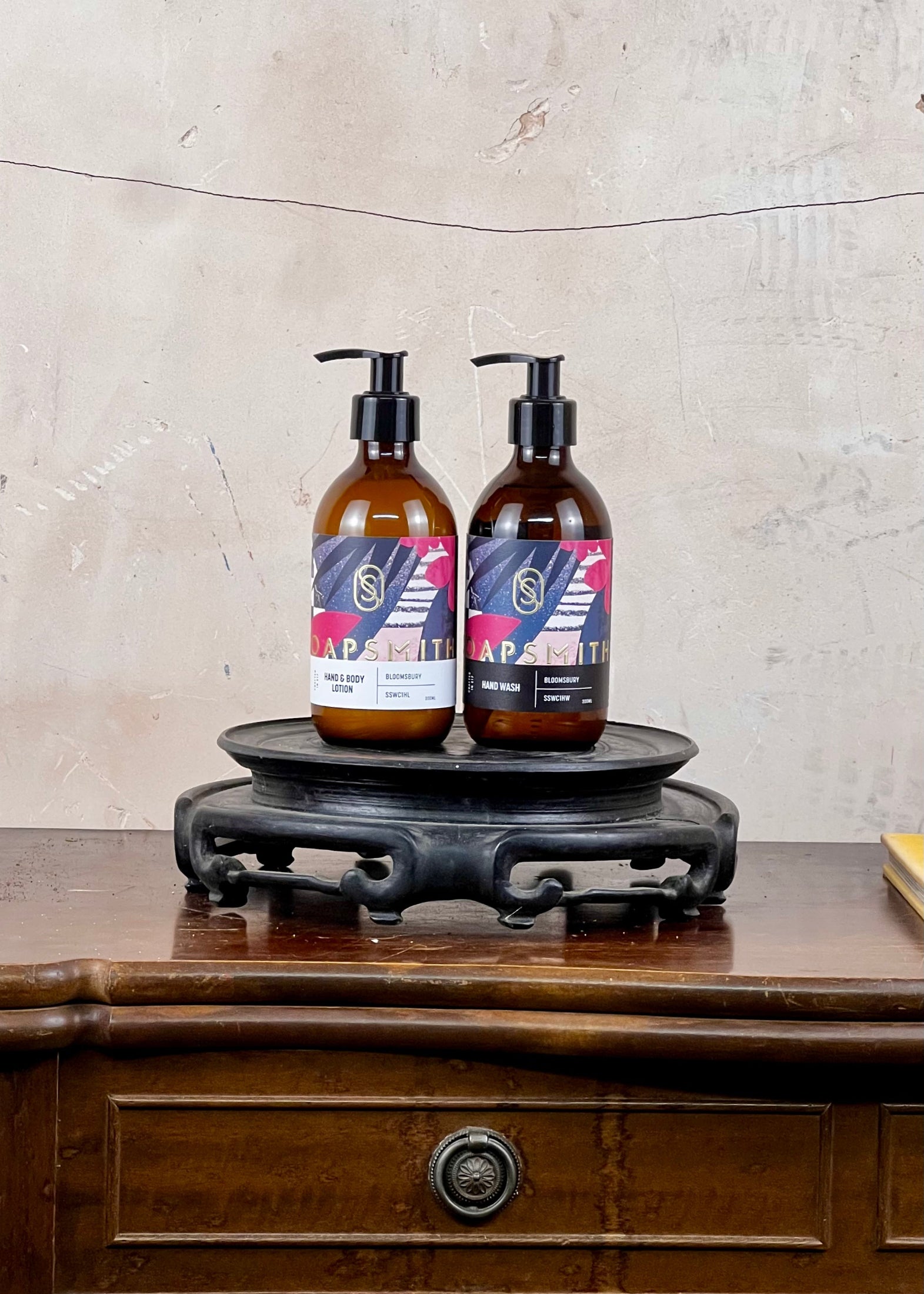 Two bottles with labels on a wooden stand against a textured wall.