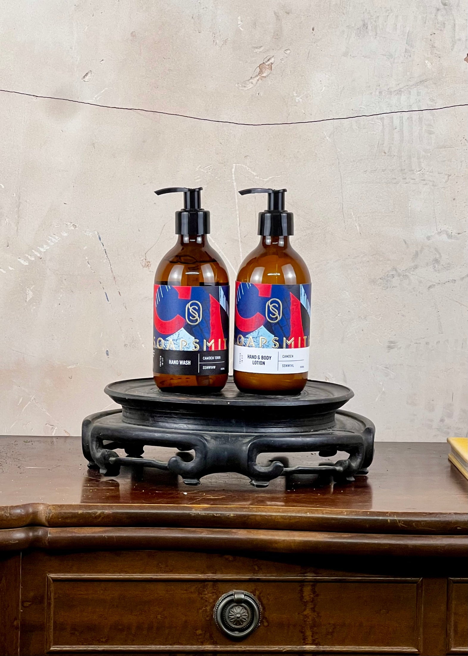 Two bottles with colorful labels on a wooden stand against a textured wall.
