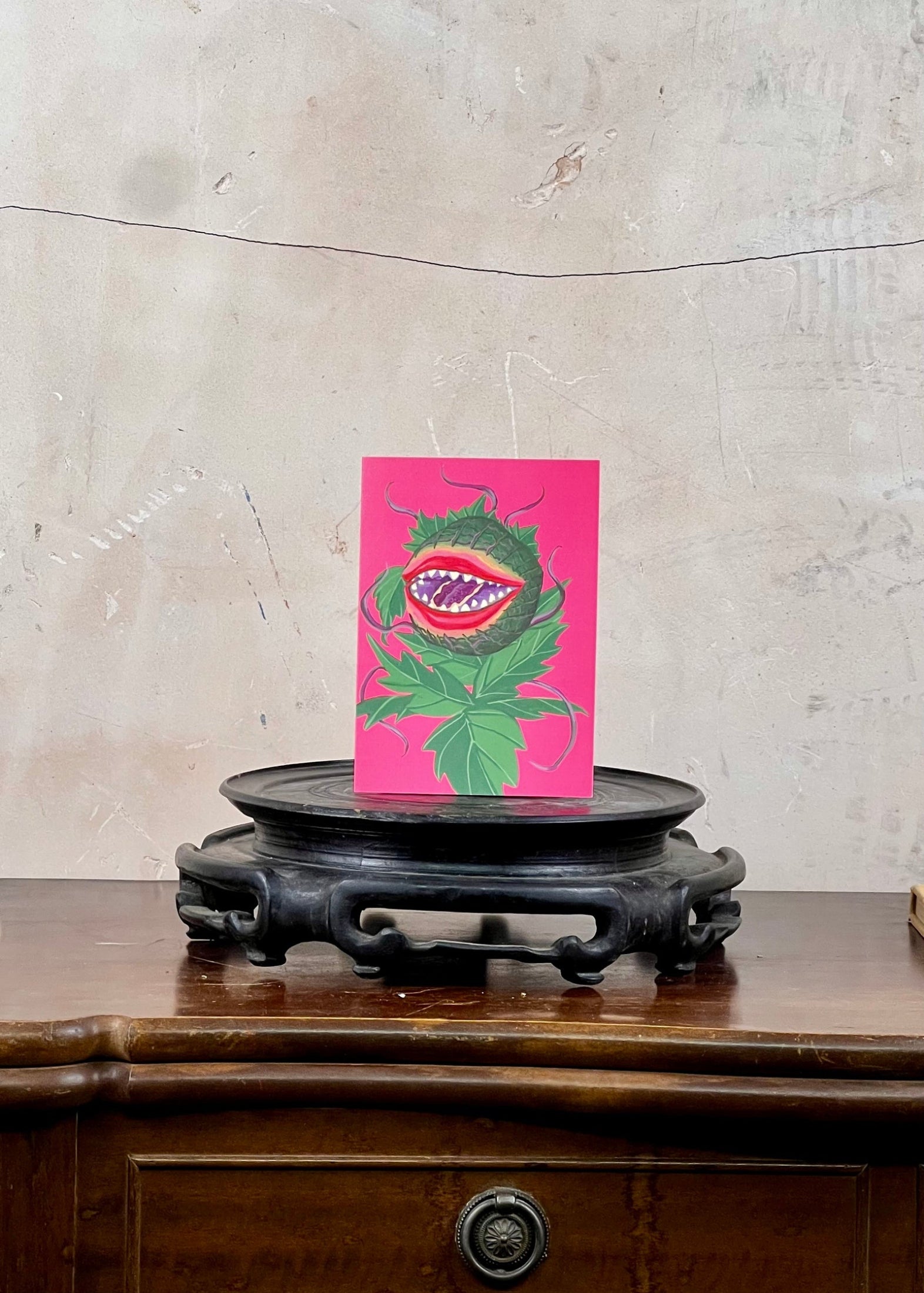 Pink card with a green creature design on a wooden surface with a textured wall background