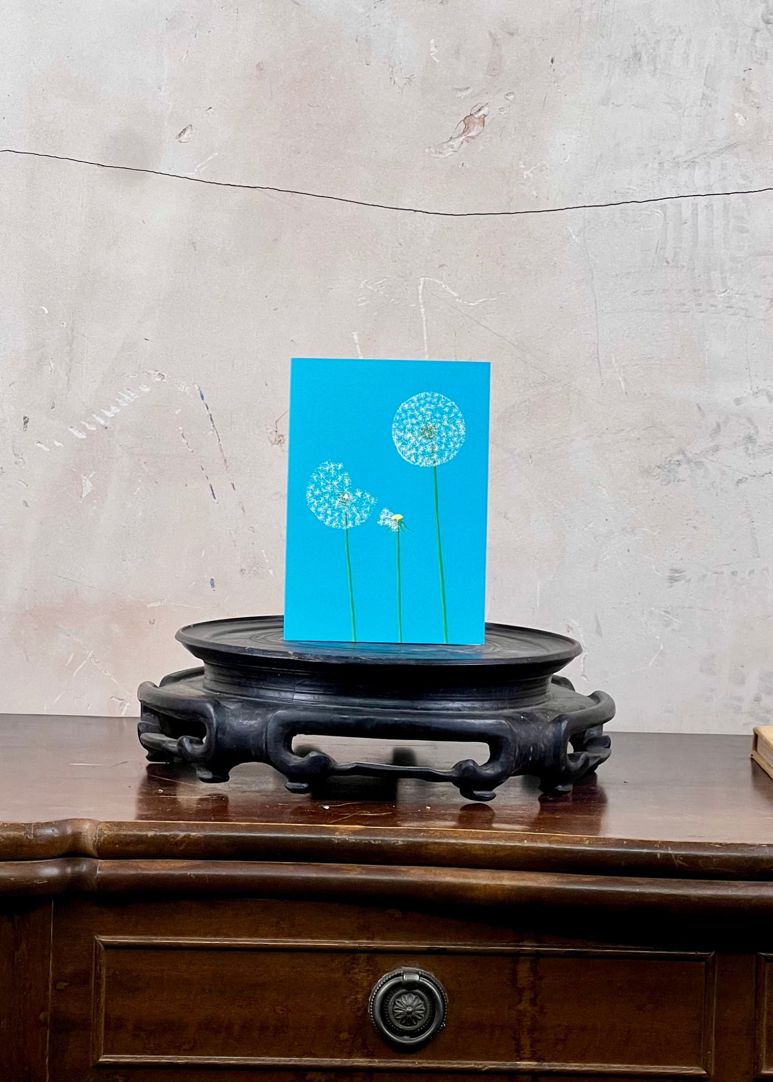 Blue card with dandelion design on a wooden stand against a concrete wall.