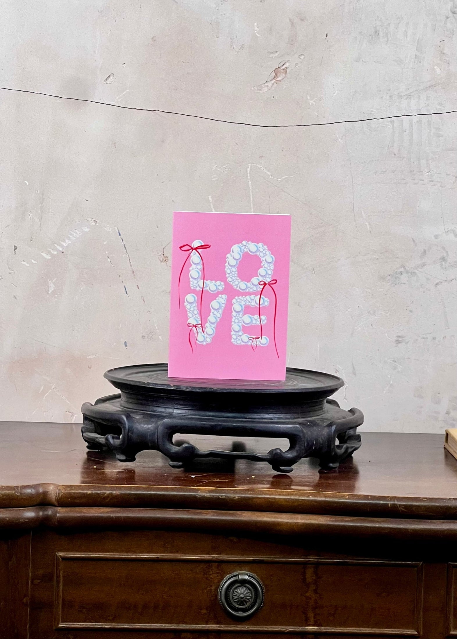 Pink card with 'LOVE' and heart designs on a wooden surface with a textured wall background