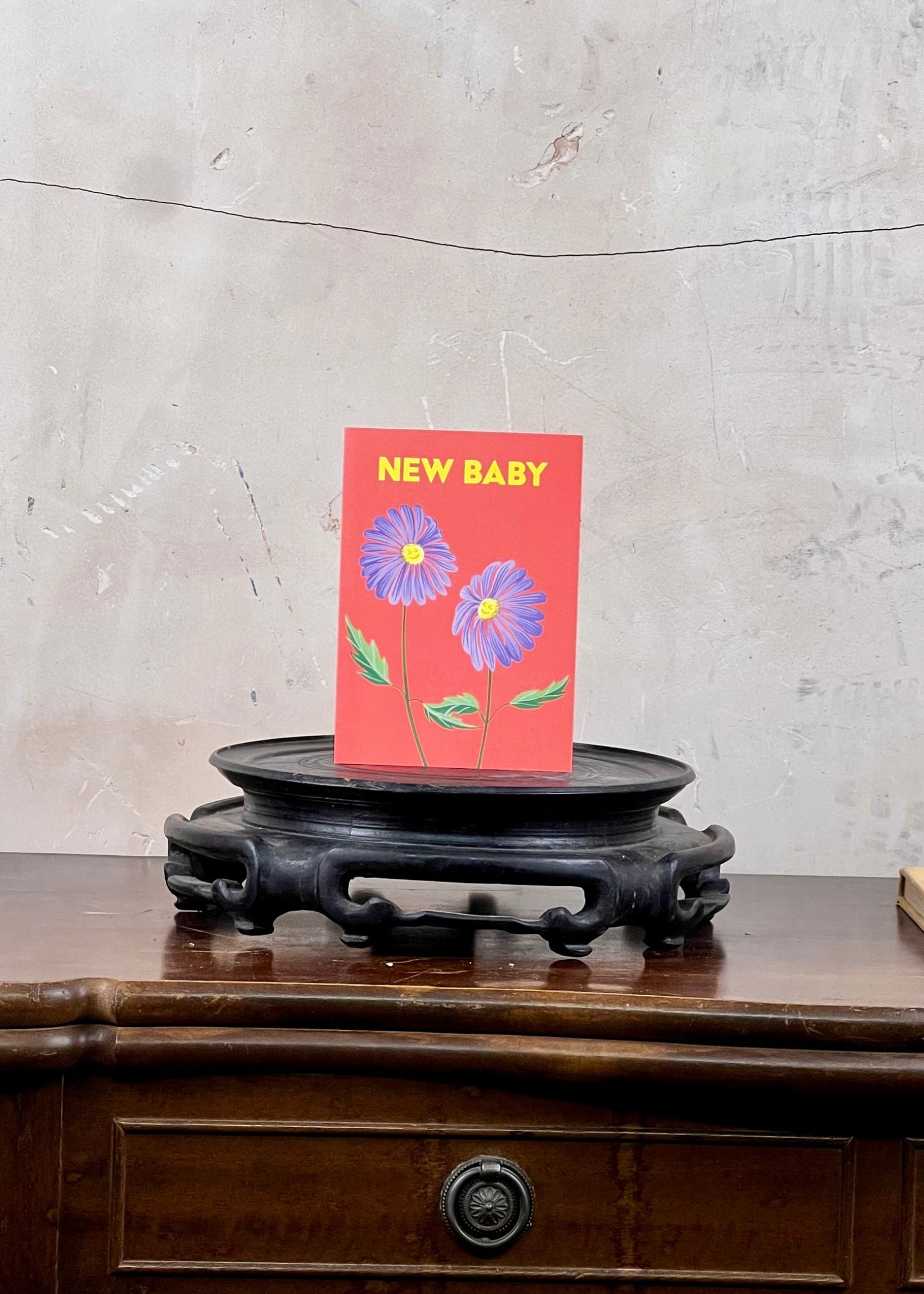 Card with flowers and 'New Baby' text on a wooden stand against a concrete wall.