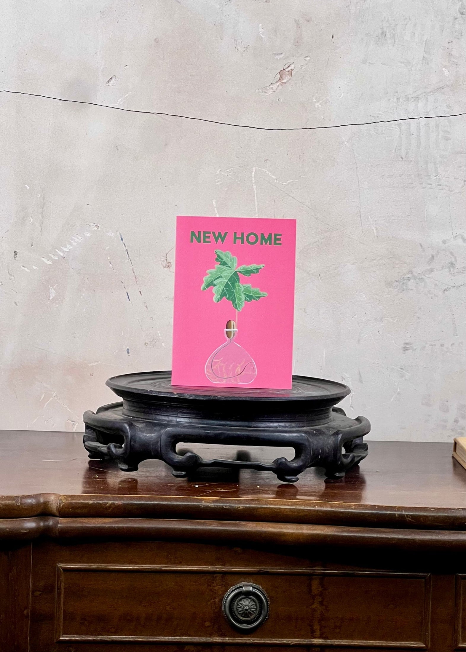 Pink 'New Home' card with a green leaf design on a wooden surface against a concrete wall.