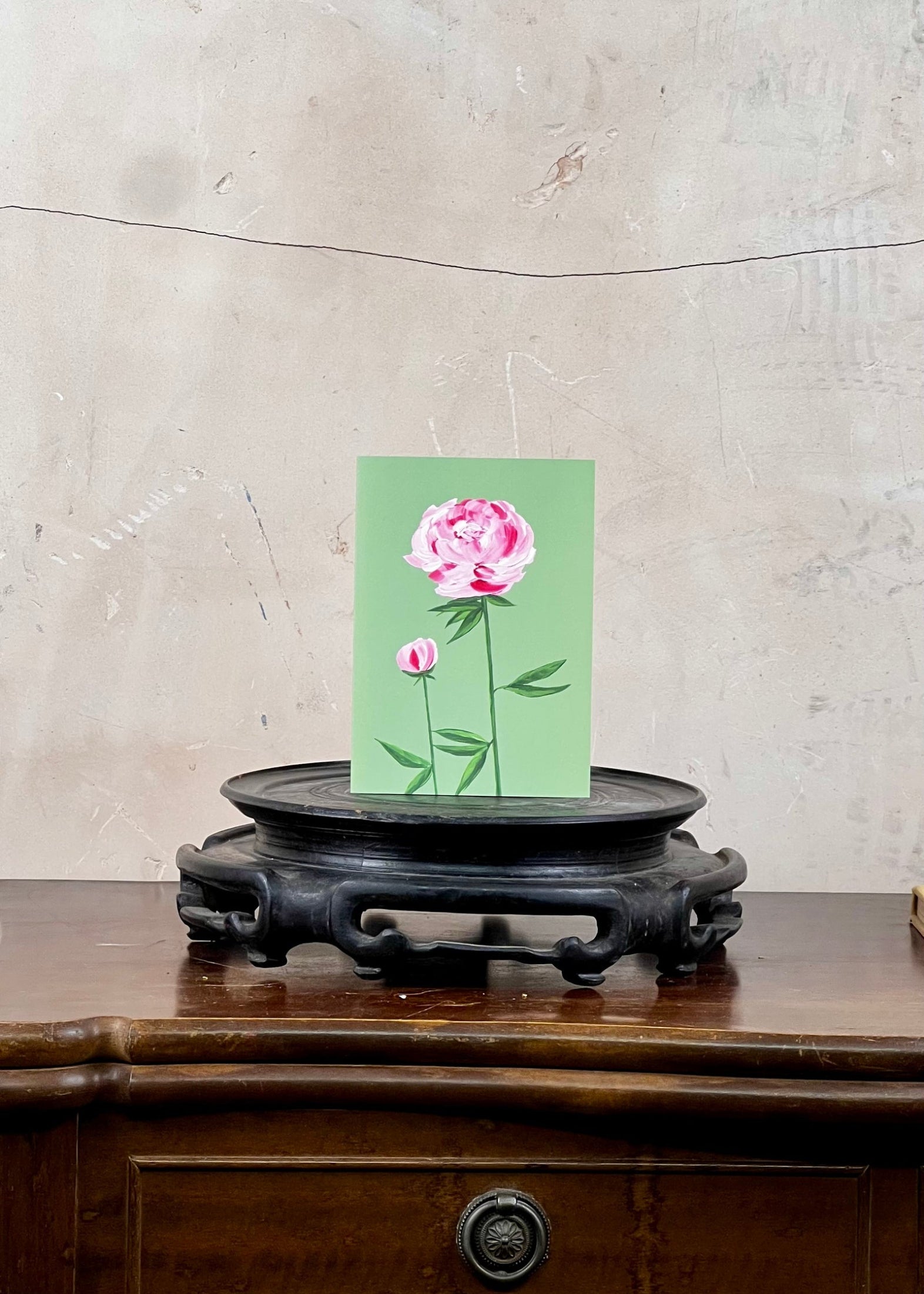 Card with pink floral design on a green background, placed on a wooden surface with a textured wall in the background.