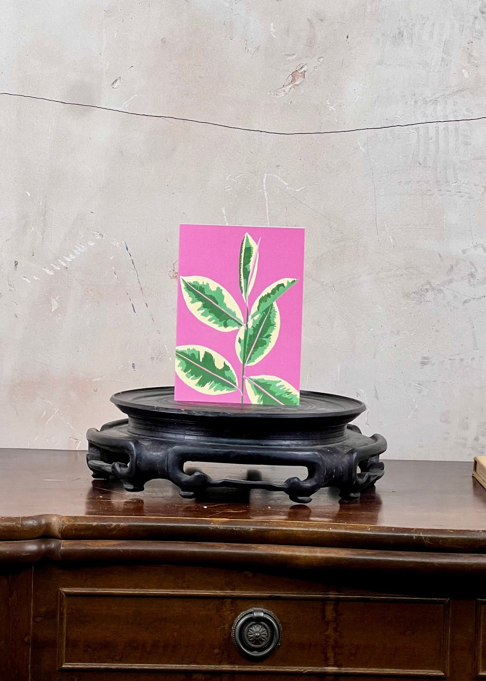 Card with green leaf design on pink background on a wooden stand against a textured wall.