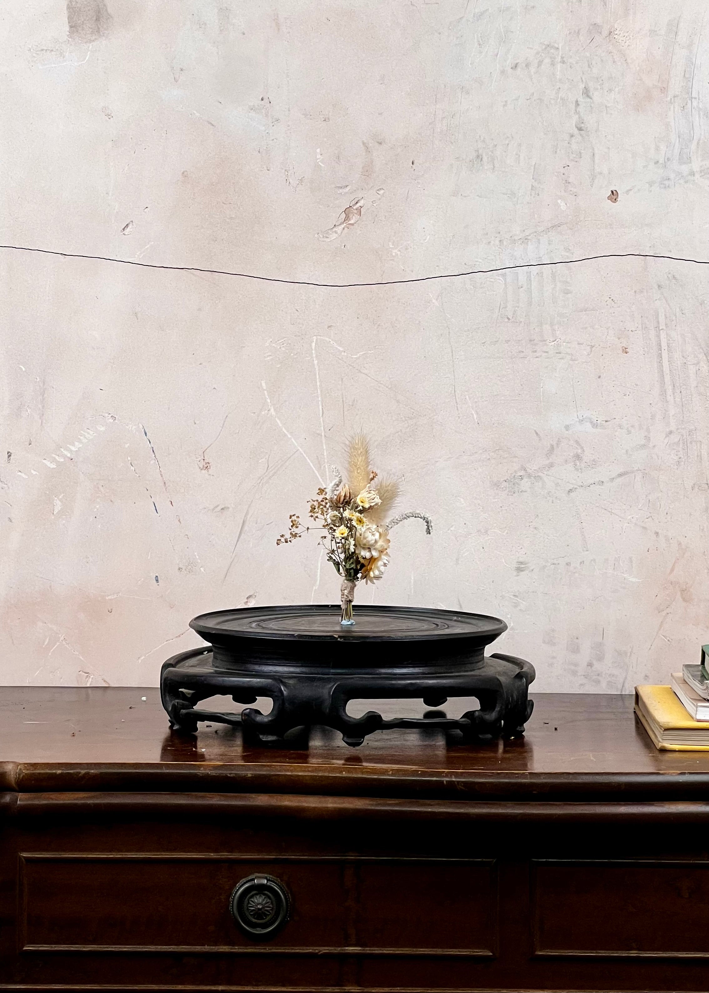 Dried flower buttonhole on a dark wooden stand against a textured white wall.