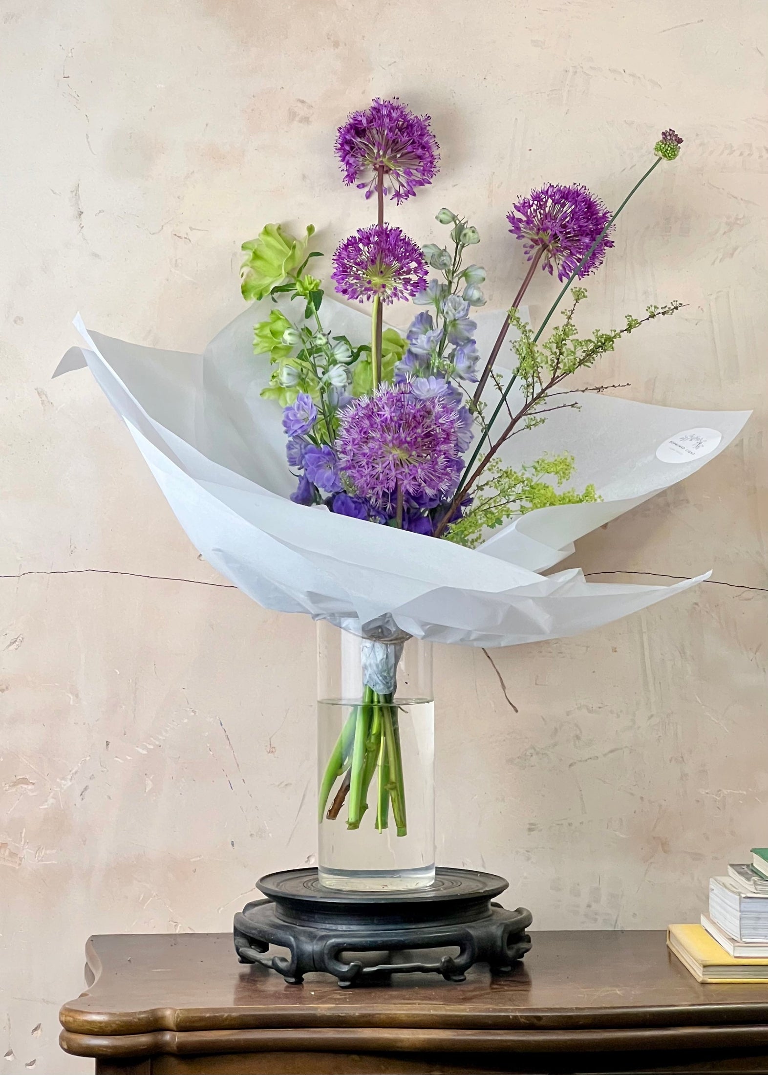 Bouquet of purple and green flowers in a clear vase on a wooden table with a beige wall background.