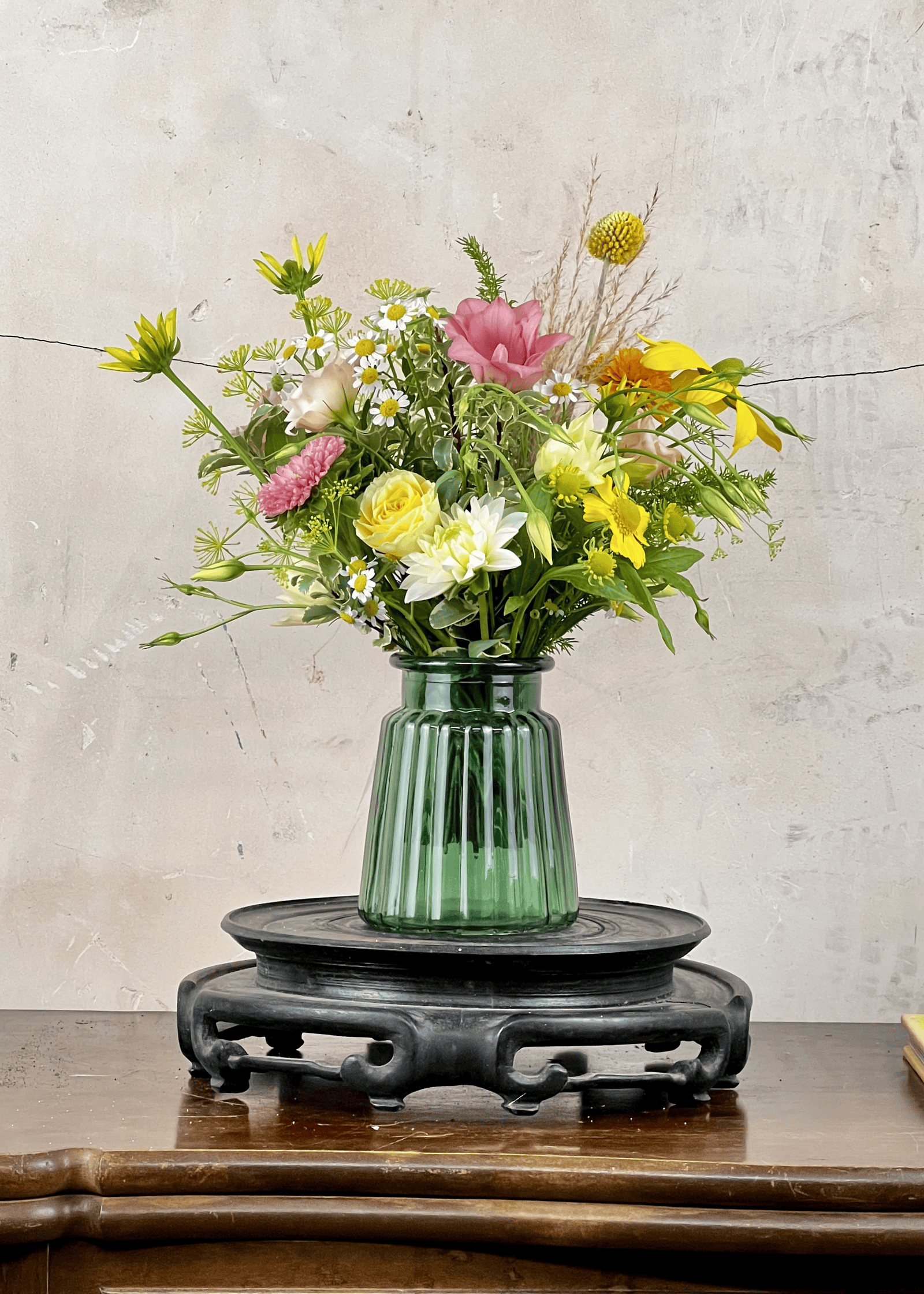 Green vase with a bouquet of flowers on a wooden table against a textured wall.