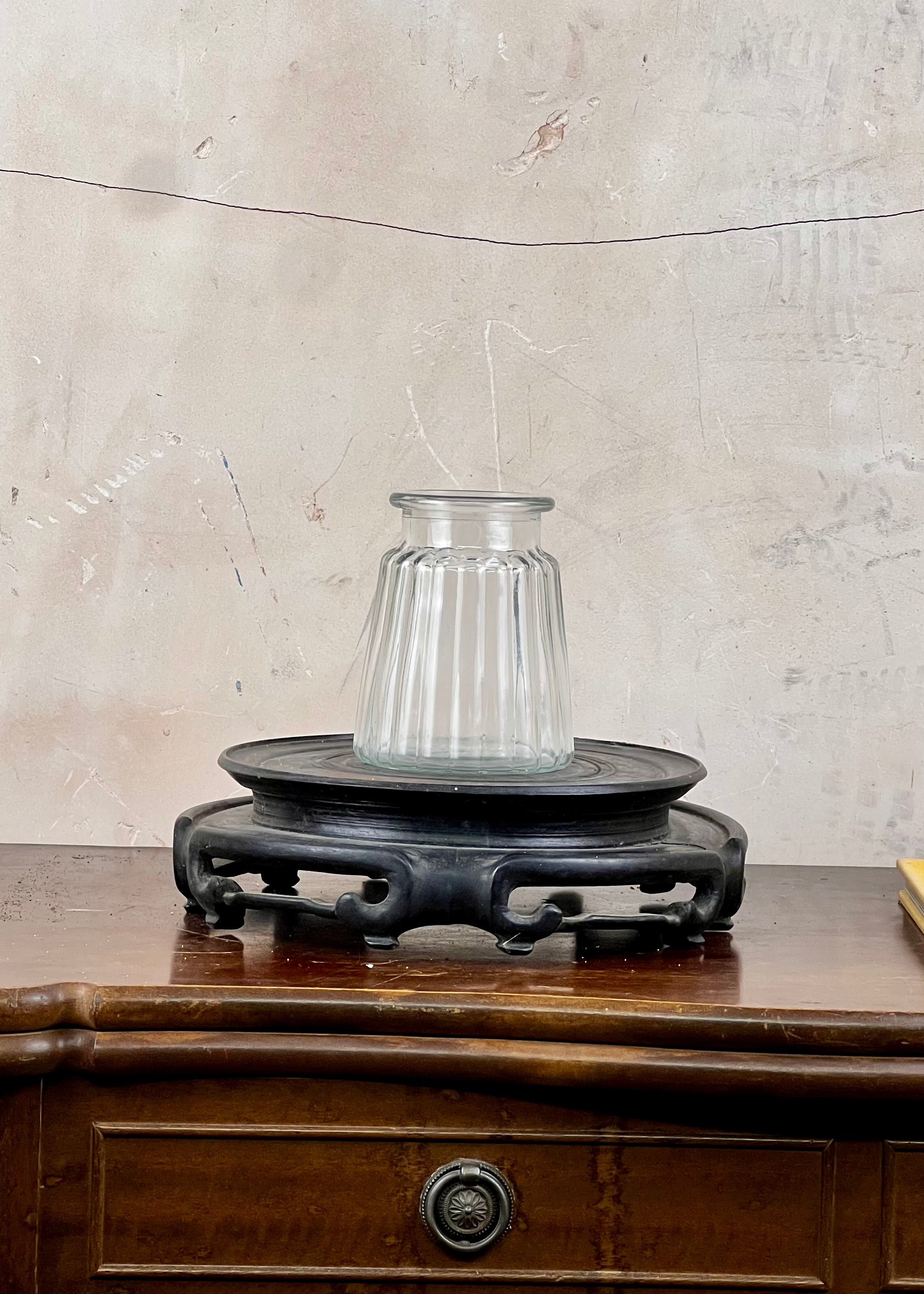 Clear glass vase on a dark wooden stand against a textured beige wall.