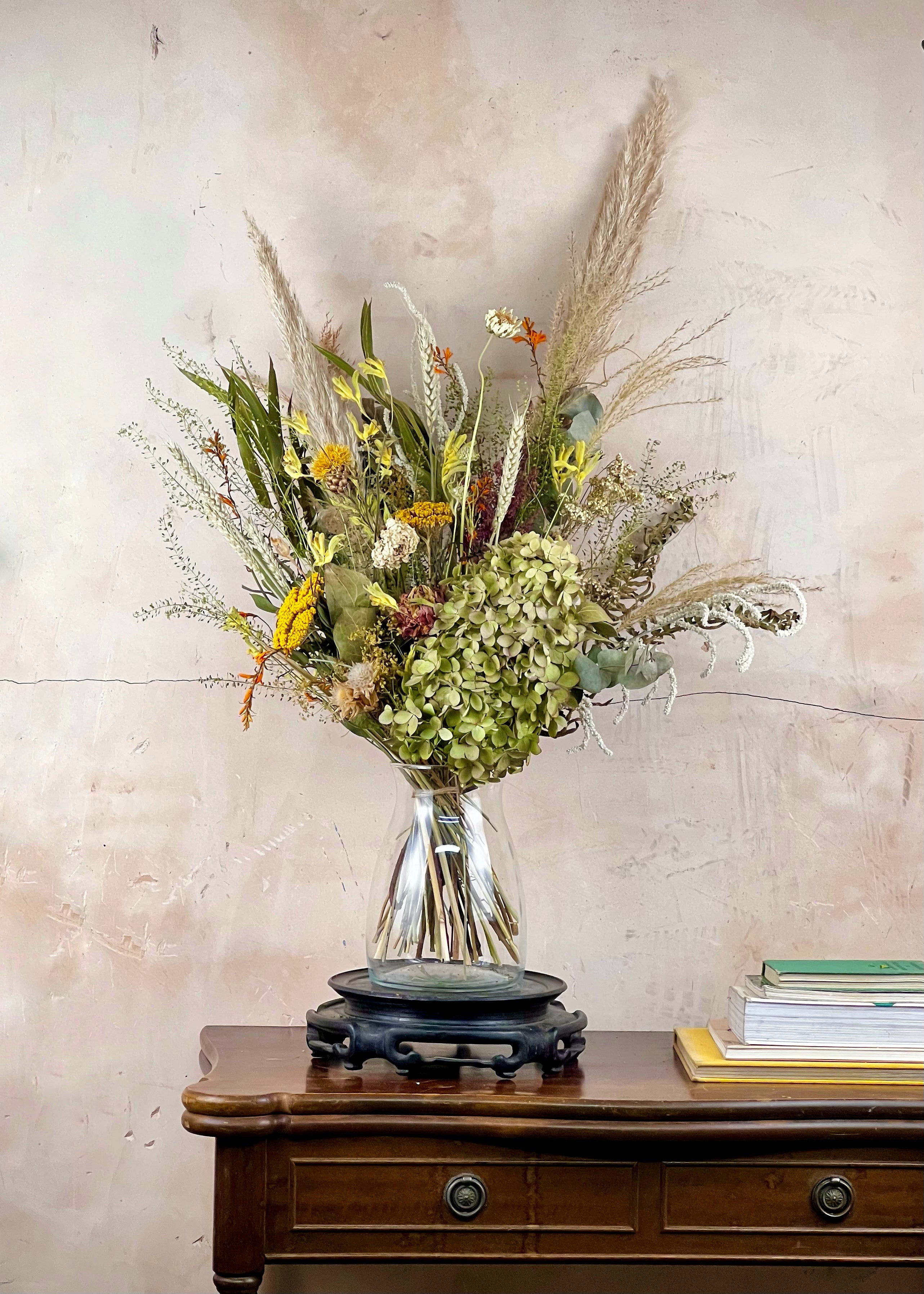 Bouquet of dried flowers in a clear vase on a wooden table against a beige wall.