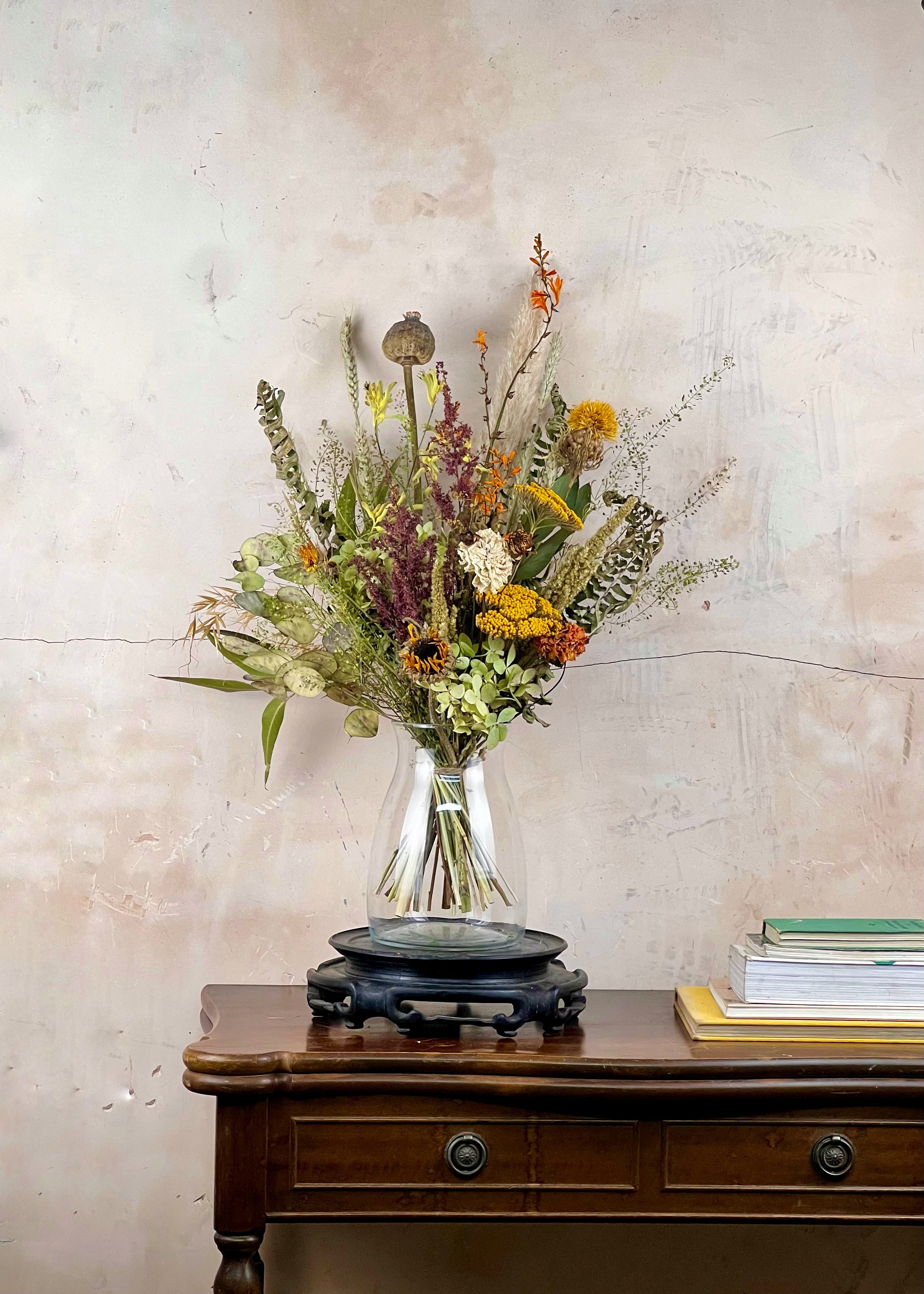 Bouquet of dried wildflowers in a clear vase on a wooden table against a textured wall.