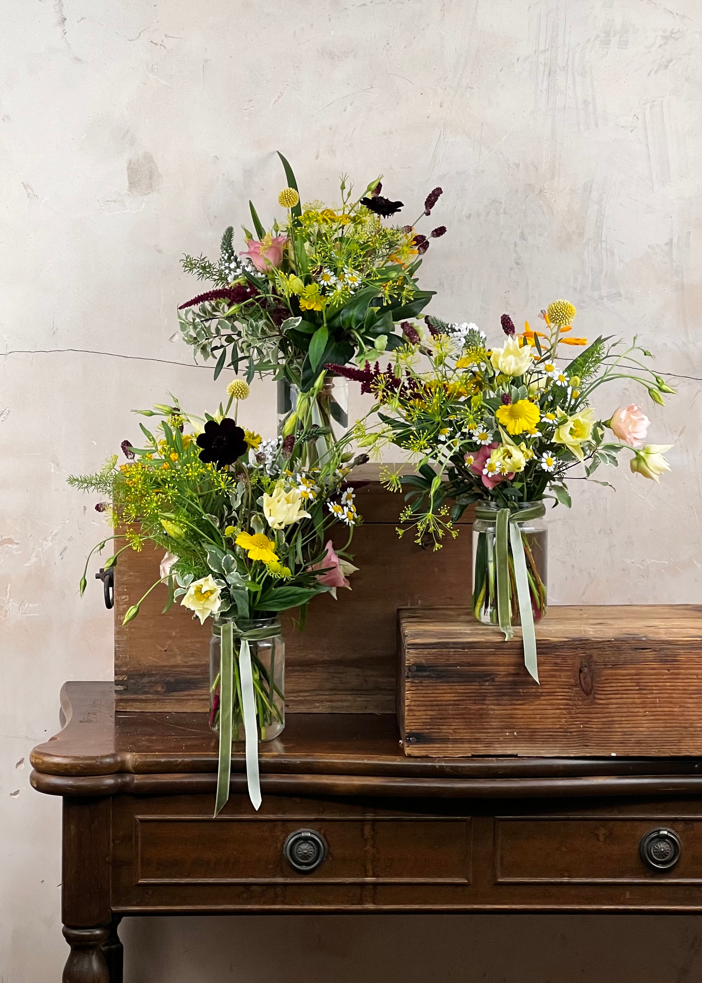 Three floral arrangements in glass jam jar vases on a wooden surface with a textured wall background.