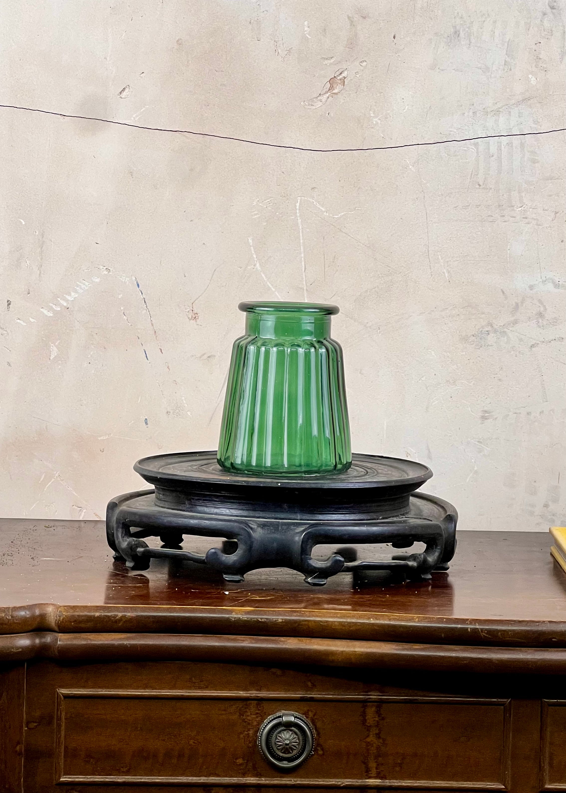 Green glass vase on a wooden stand against a textured wall.