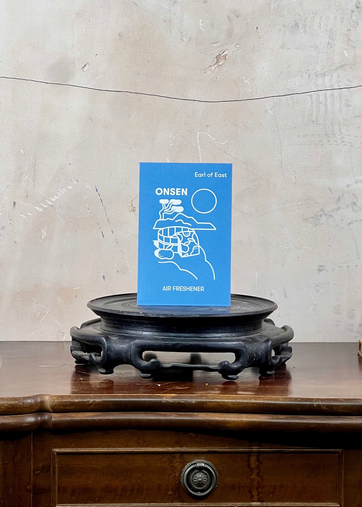 Blue air freshener titled 'Onsen' on a wooden stand against a textured wall.