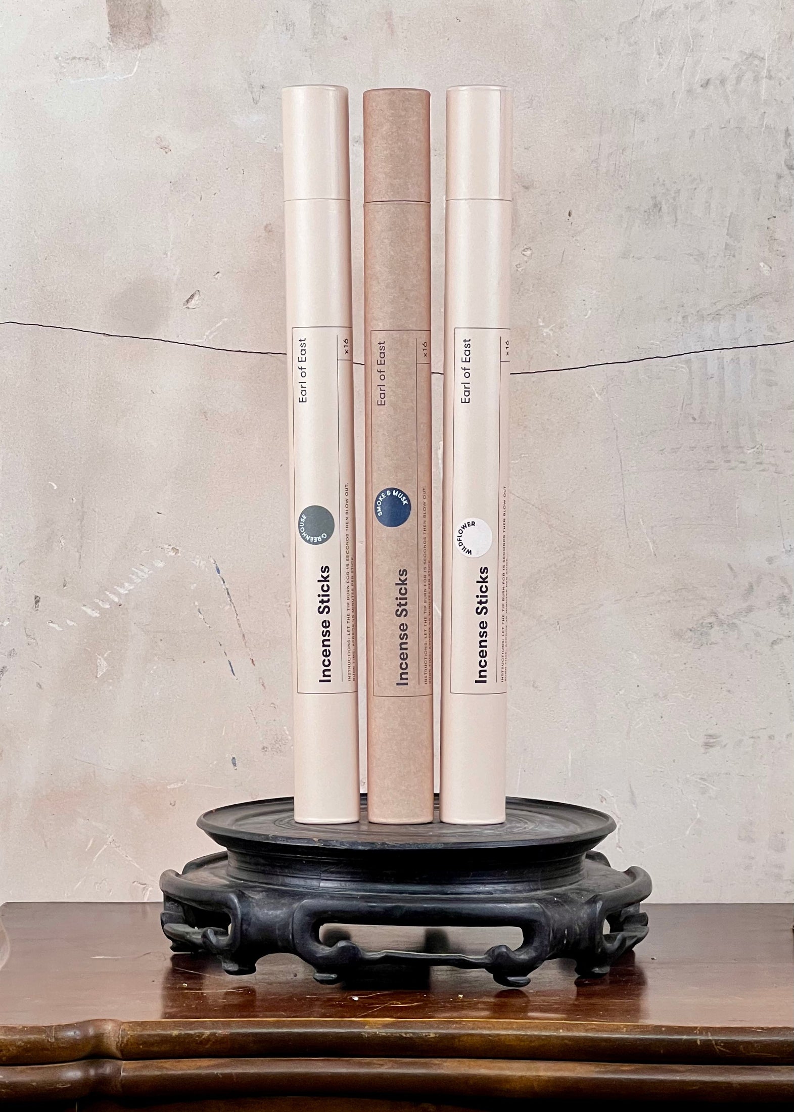 Three cylindrical incense stick containers on a dark wooden stand against a textured light gray wall.