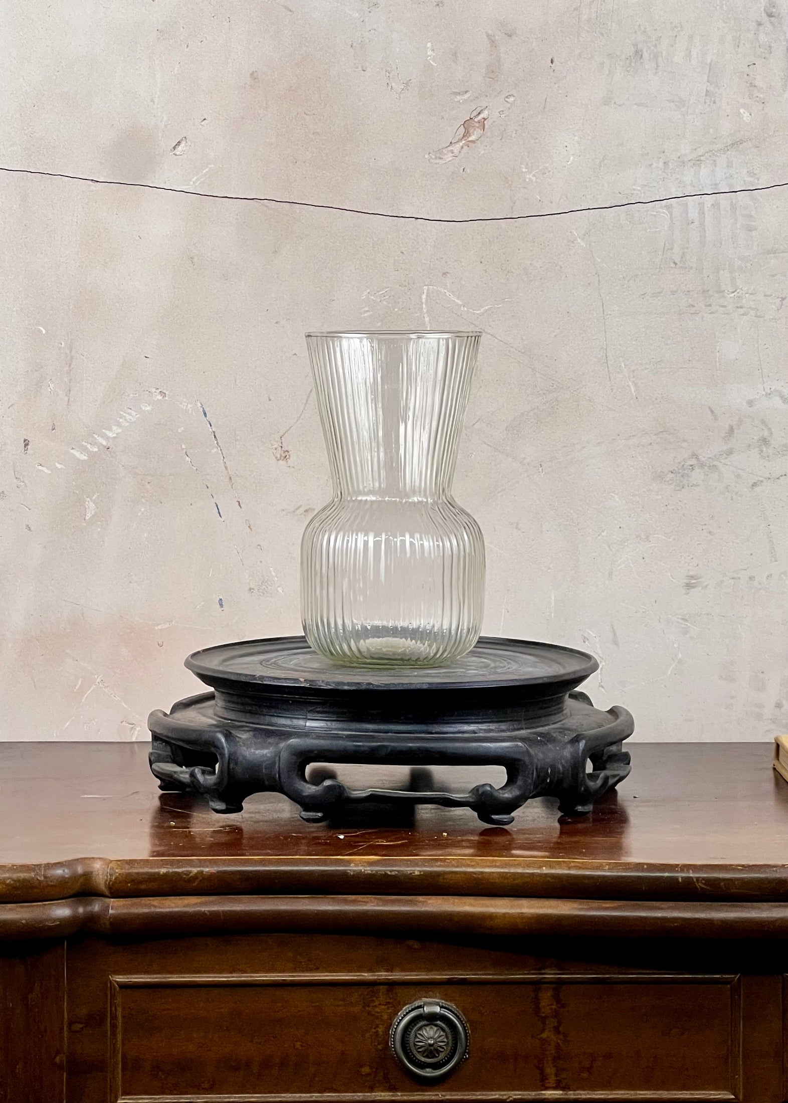 Clear ribbed glass vase on a wooden stand against a textured wall.