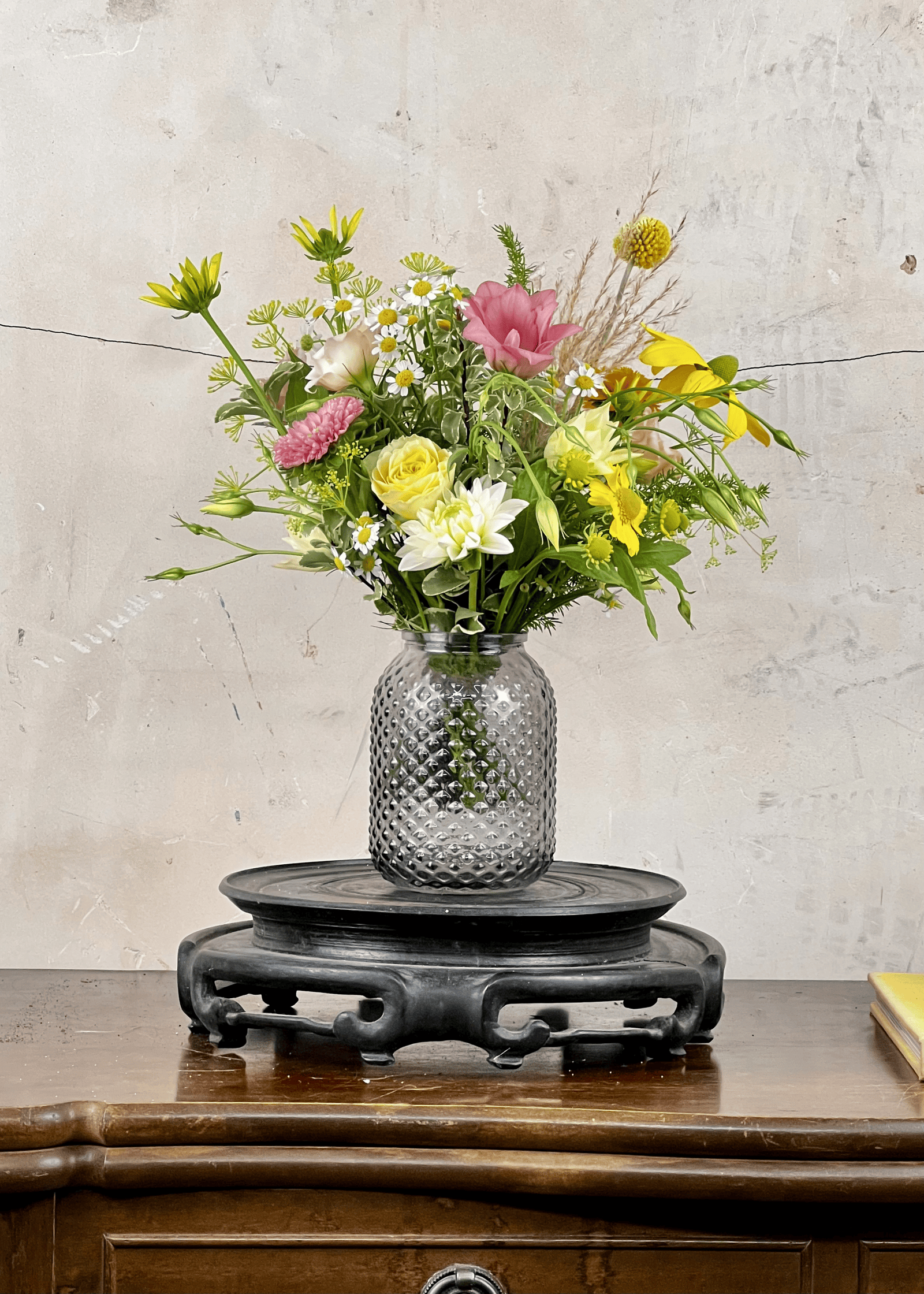 Grey clear glass vase with a bouquet of flowers on a wooden table against a textured wall.