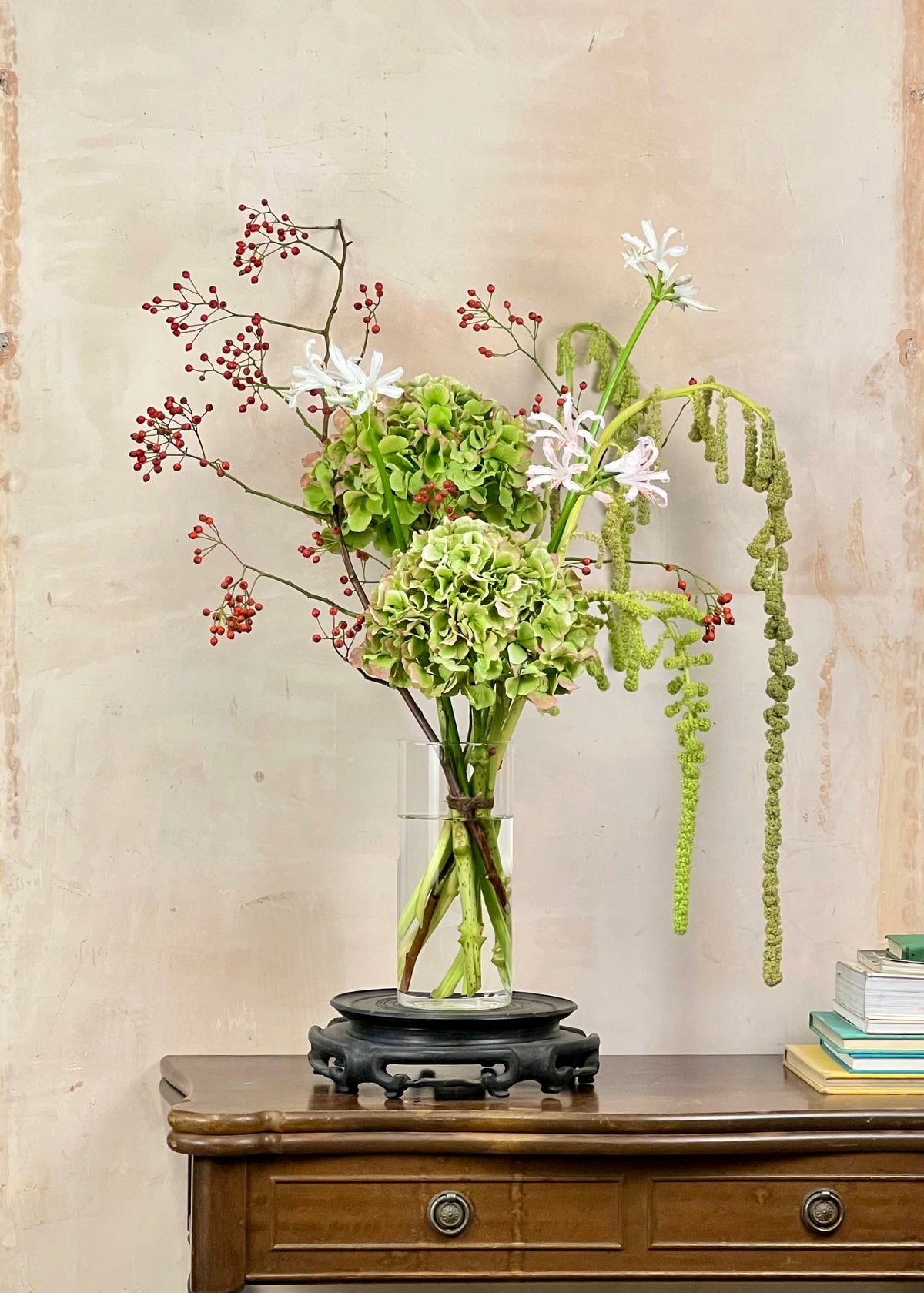 A floral arrangement in a clear glass vase with a black wooden base, displayed on a wooden cabinet against a textured beige wall. The bouquet includes green hydrangea-like blooms, white flowers, red berries, and trailing green stems.