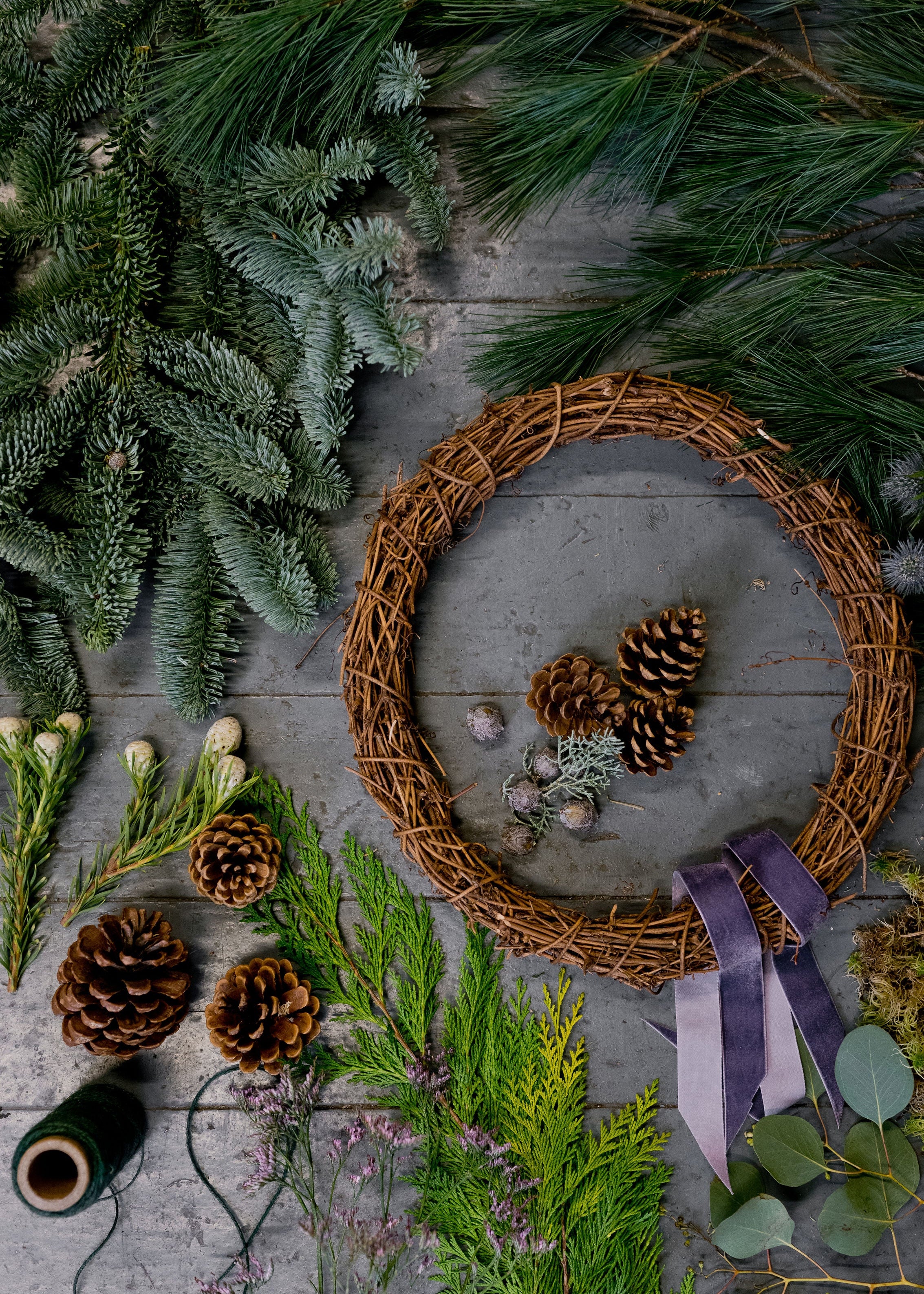 Wreath with pine cones and ribbons on a wooden surface surrounded by greenery