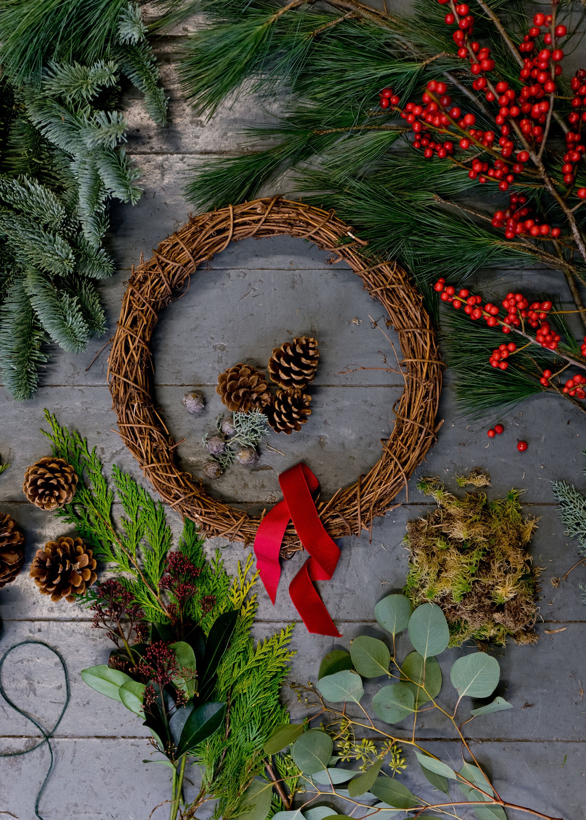 Wreath with red ribbon surrounded by pinecones, greenery, and berries on a wooden surface