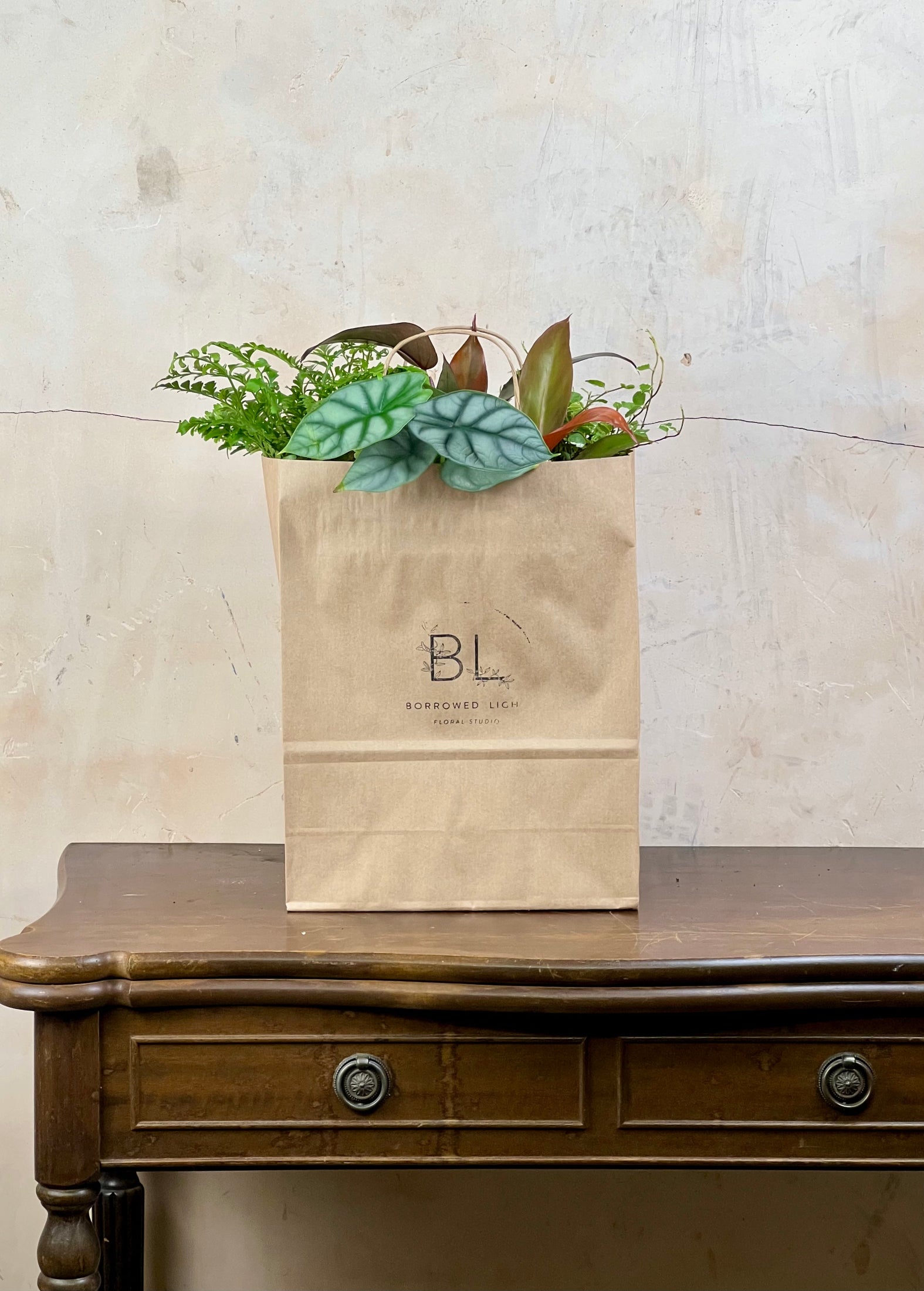 A kraft paper gift bag with the logo 'BL' and 'BLOOMBERG LUXE' printed on the front, filled with green and brown artificial foliage, sitting on a dark wood console table against a textured beige wall.